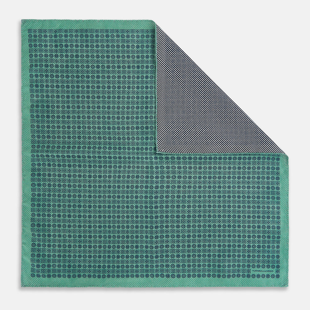 A square silk pocket square in Rosette Green by Turnbull & Asser UK, featuring a teal base with a grid of dark dots. One corner is folded to showcase the black and white microdot pattern on the reverse, while subtle hints of navy, aqua, and green form geometric medallions that lend a refined modern touch.