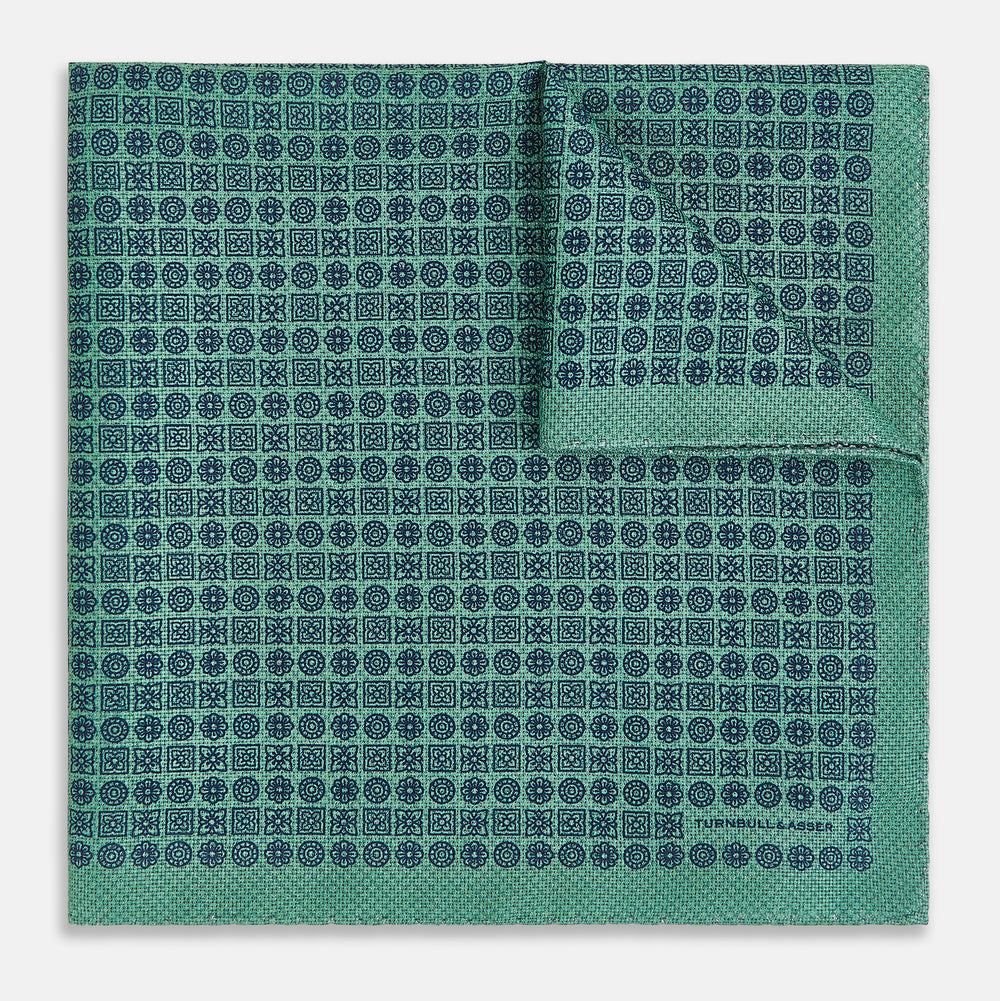 A square silk pocket square in Rosette Green by Turnbull & Asser UK, featuring a teal base with a grid of dark dots. One corner is folded to showcase the black and white microdot pattern on the reverse, while subtle hints of navy, aqua, and green form geometric medallions that lend a refined modern touch.