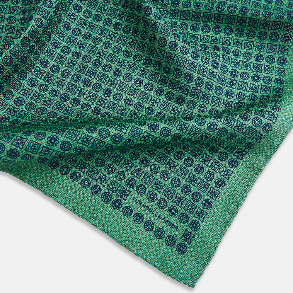 A square silk pocket square in Rosette Green by Turnbull & Asser UK, featuring a teal base with a grid of dark dots. One corner is folded to showcase the black and white microdot pattern on the reverse, while subtle hints of navy, aqua, and green form geometric medallions that lend a refined modern touch.