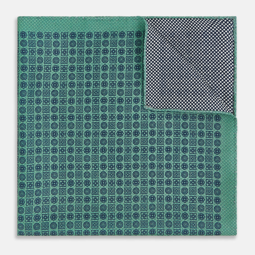 A square silk pocket square in Rosette Green by Turnbull & Asser UK, featuring a teal base with a grid of dark dots. One corner is folded to showcase the black and white microdot pattern on the reverse, while subtle hints of navy, aqua, and green form geometric medallions that lend a refined modern touch.