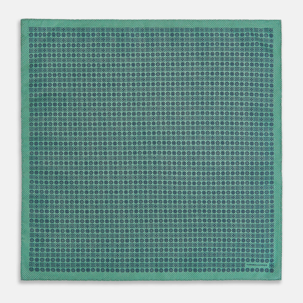 A square silk pocket square in Rosette Green by Turnbull & Asser UK, featuring a teal base with a grid of dark dots. One corner is folded to showcase the black and white microdot pattern on the reverse, while subtle hints of navy, aqua, and green form geometric medallions that lend a refined modern touch.