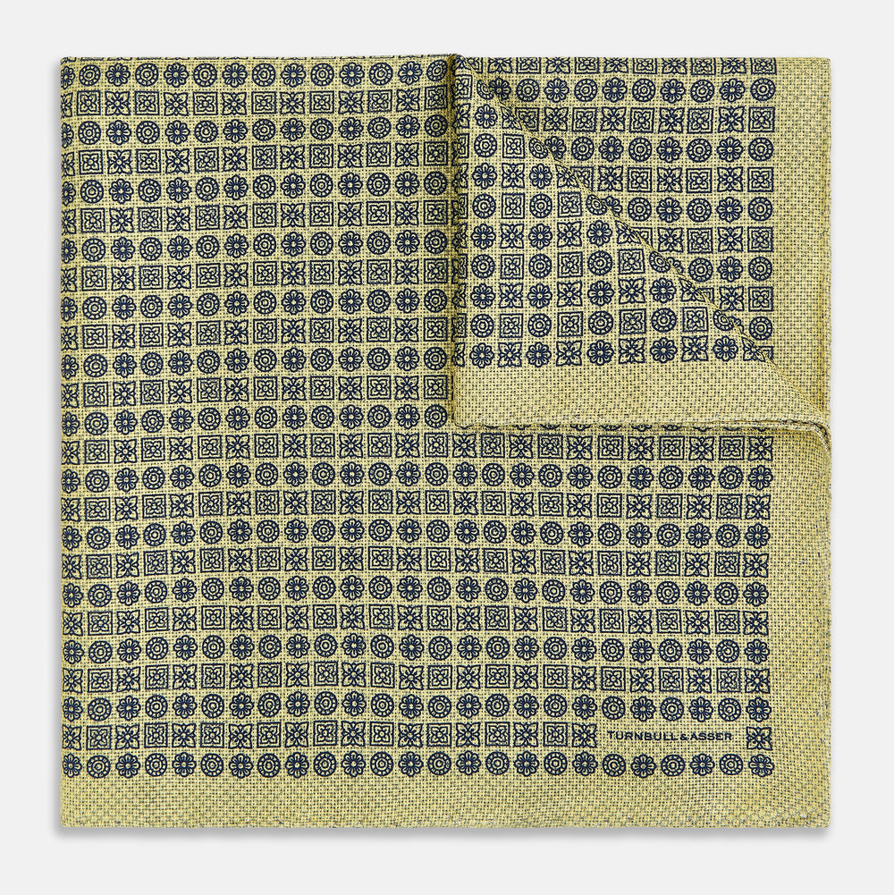 The Rosette Yellow Pocket Square in Silk by Turnbull & Asser UK features a square design with a yellow border, adorned with a grid of small black and white circles. One corner is neatly folded to reveal a reverse side in a refined black and white micro-checkered pattern, while subtle geometric medallions lend an added touch of elegance to the overall look.