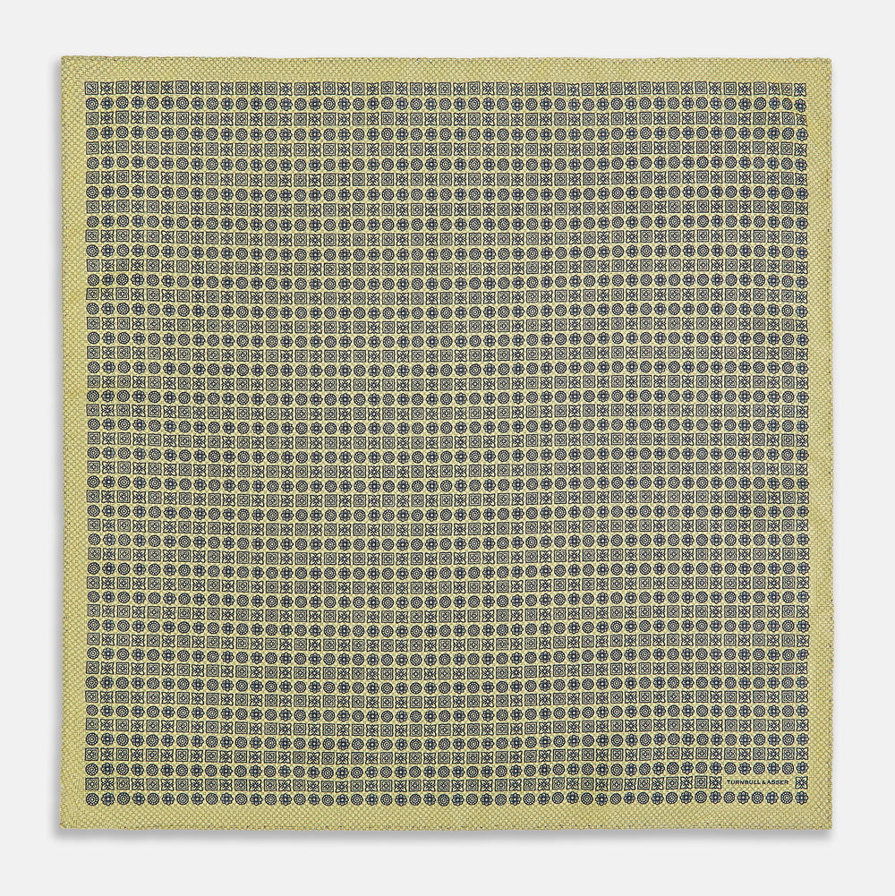 The Rosette Yellow Pocket Square in Silk by Turnbull & Asser UK features a square design with a yellow border, adorned with a grid of small black and white circles. One corner is neatly folded to reveal a reverse side in a refined black and white micro-checkered pattern, while subtle geometric medallions lend an added touch of elegance to the overall look.