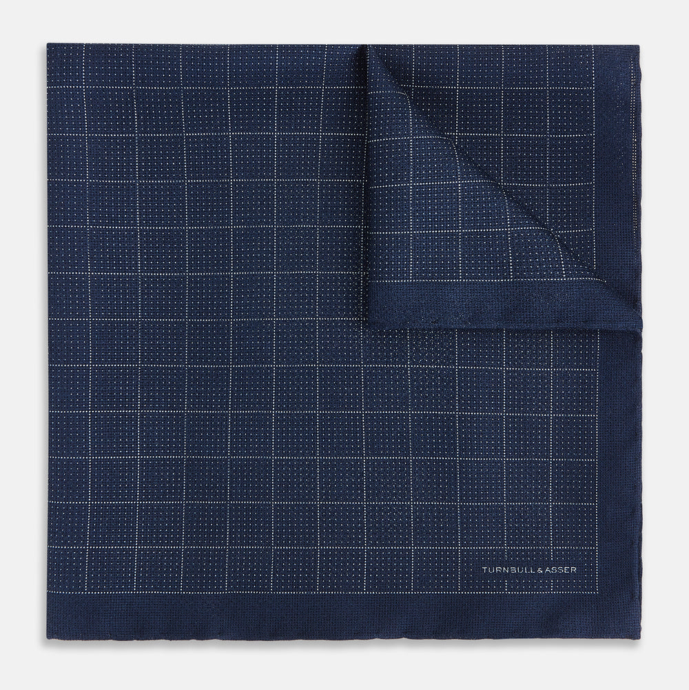 The Checked Blue Pocket Square in Silk by Turnbull & Asser UK features a navy base with a light blue grid pattern on one side and a refined dotted design on the other. This double-sided accessory is shown partially folded to elegantly showcase both distinctive patterns.