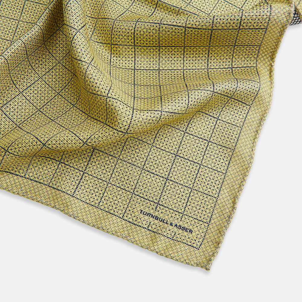 A yellow silk pocket square featuring a small black grid pattern of dotted squares, neatly folded at one corner. "Turnbull & Asser UK" is printed in the bottom right corner.