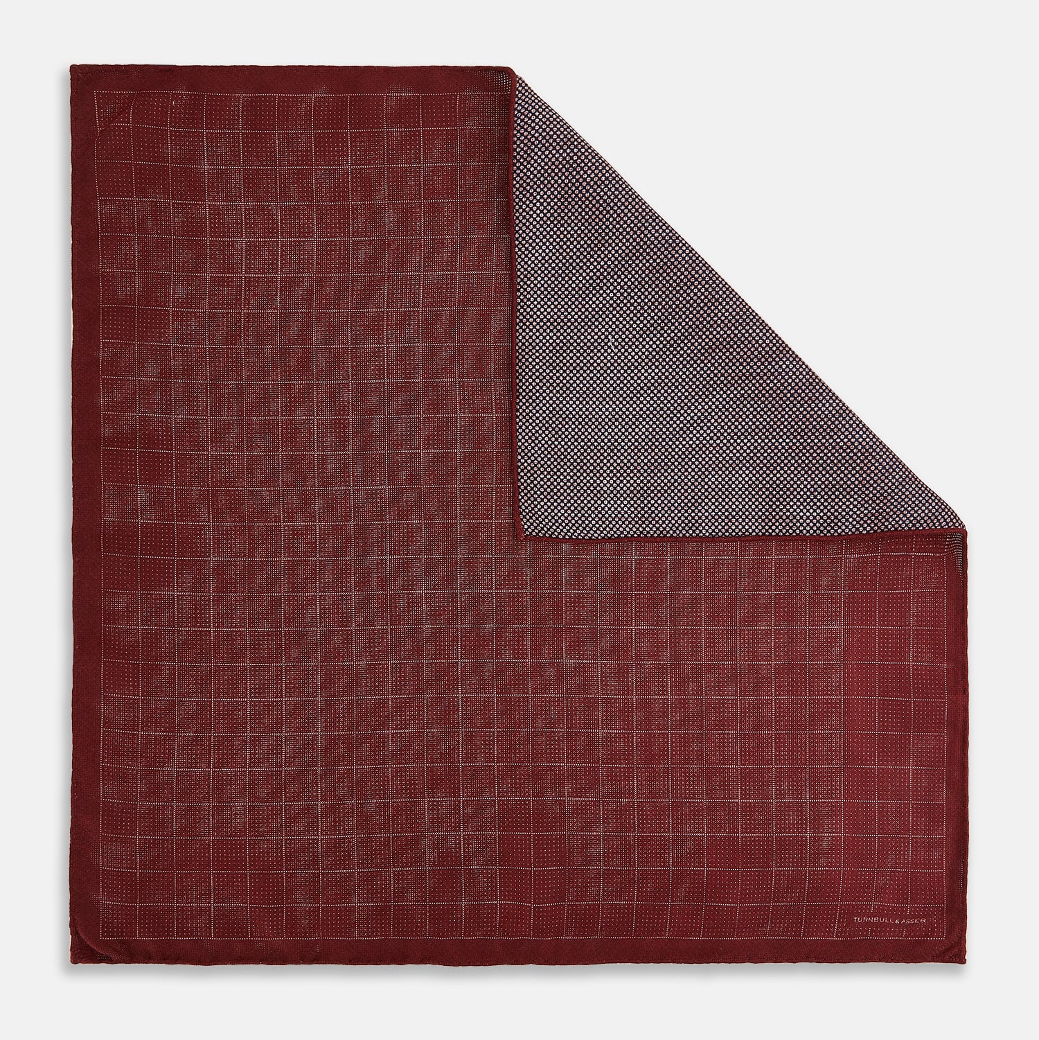View 1 of product The Turnbull & Asser UK Checked Burgundy Pocket Square in Silk features a sophisticated burgundy hue with a subtle grid pattern. It's displayed folded at one corner to reveal a contrasting small checkered design on the reverse, showcasing its refined double-sided craftsmanship.