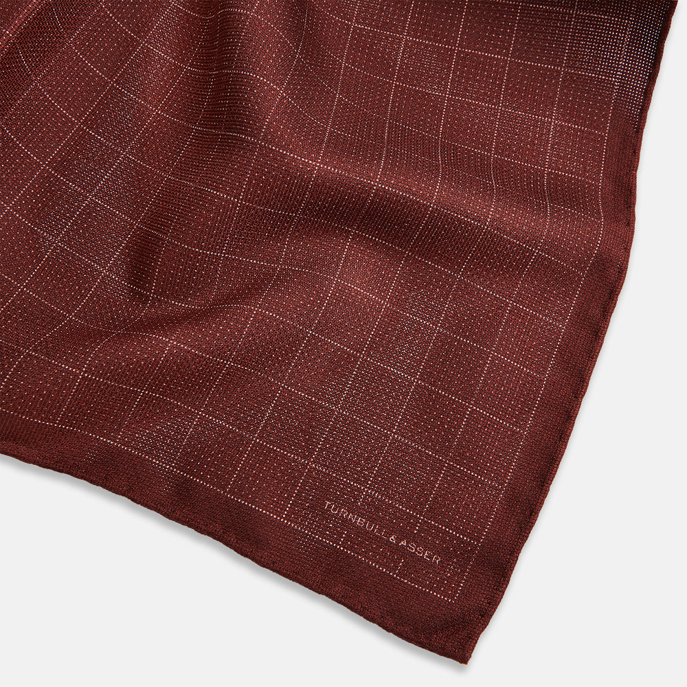 The Turnbull & Asser UK Checked Burgundy Pocket Square in Silk features a sophisticated burgundy hue with a subtle grid pattern. It's displayed folded at one corner to reveal a contrasting small checkered design on the reverse, showcasing its refined double-sided craftsmanship.
