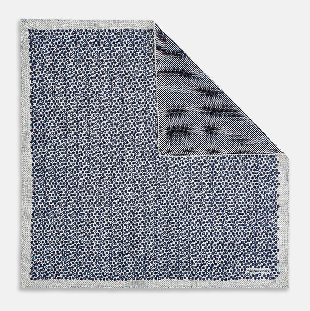 A square silk pocket square from Turnbull & Asser UK, featuring a subtle floral grey pattern; one corner is folded to reveal its double-sided design with a complementary pattern on the reverse side.