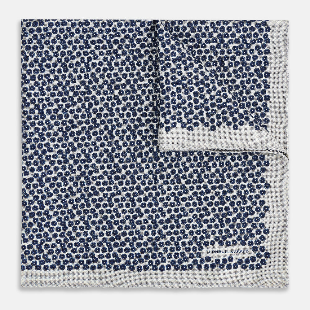 A square silk pocket square from Turnbull & Asser UK, featuring a subtle floral grey pattern; one corner is folded to reveal its double-sided design with a complementary pattern on the reverse side.