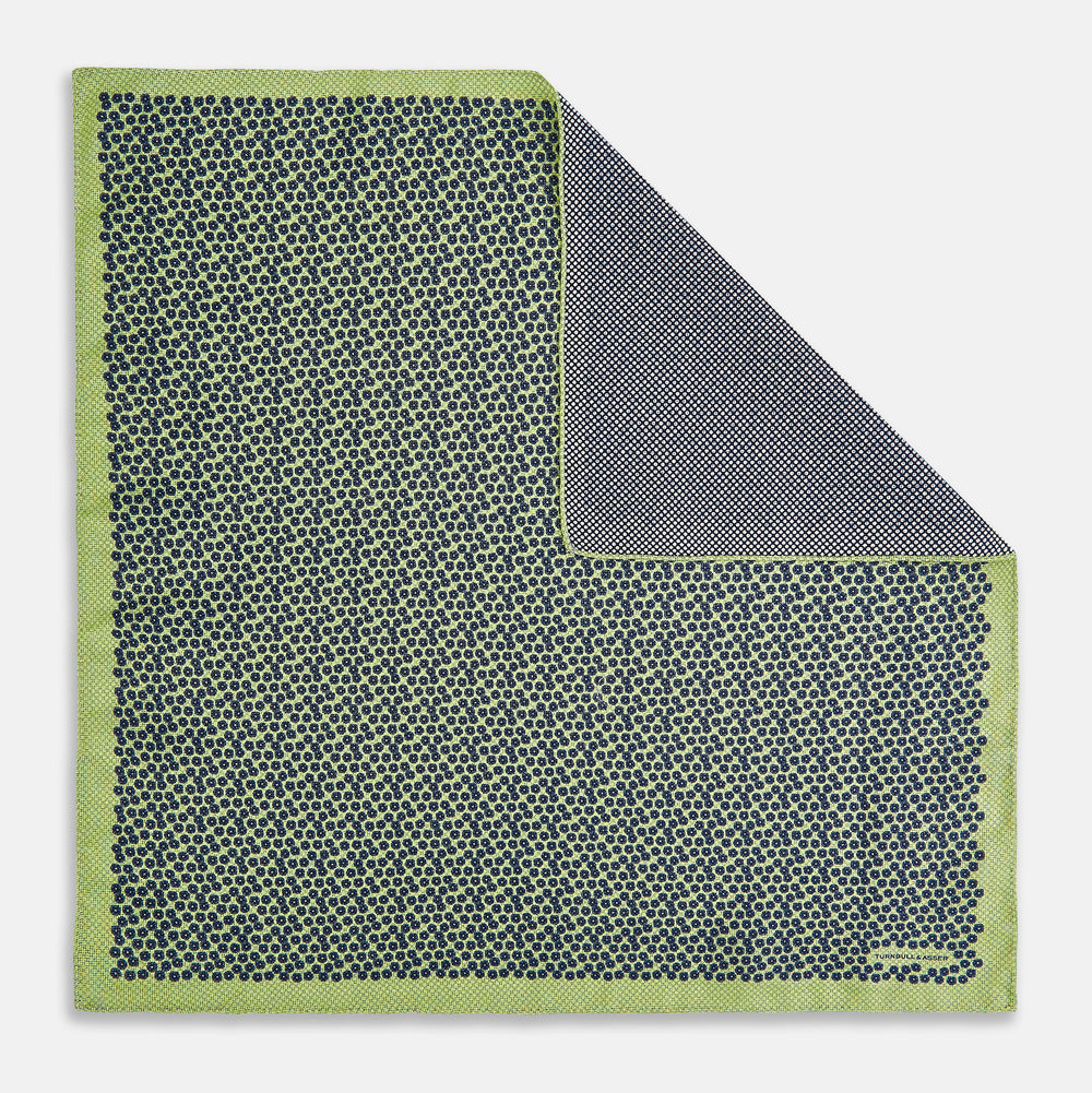 The Turnbull & Asser UK Floral Green Pocket Square in Silk is a square accessory featuring a blue floral pattern with a green border, partly folded at one corner to showcase its double-sided design, revealing a navy and light green dotted pattern on the reverse.