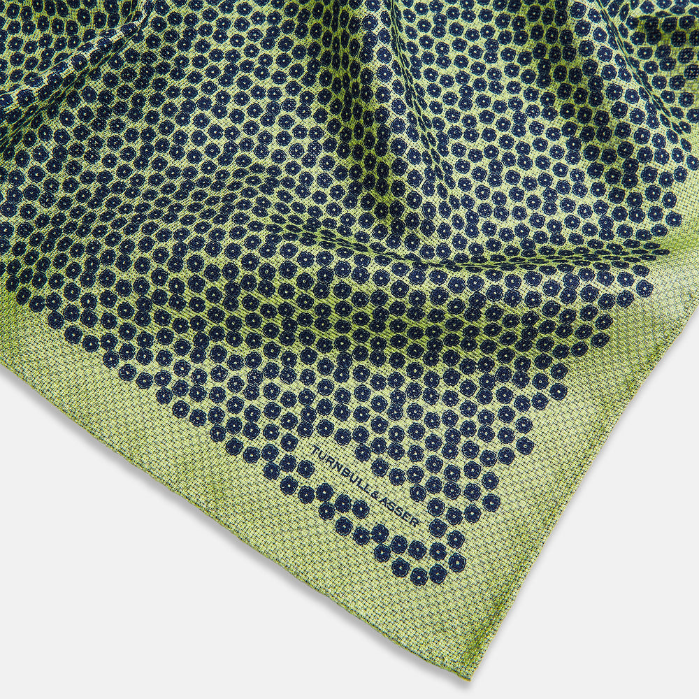 The Turnbull & Asser UK Floral Green Pocket Square in Silk is a square accessory featuring a blue floral pattern with a green border, partly folded at one corner to showcase its double-sided design, revealing a navy and light green dotted pattern on the reverse.