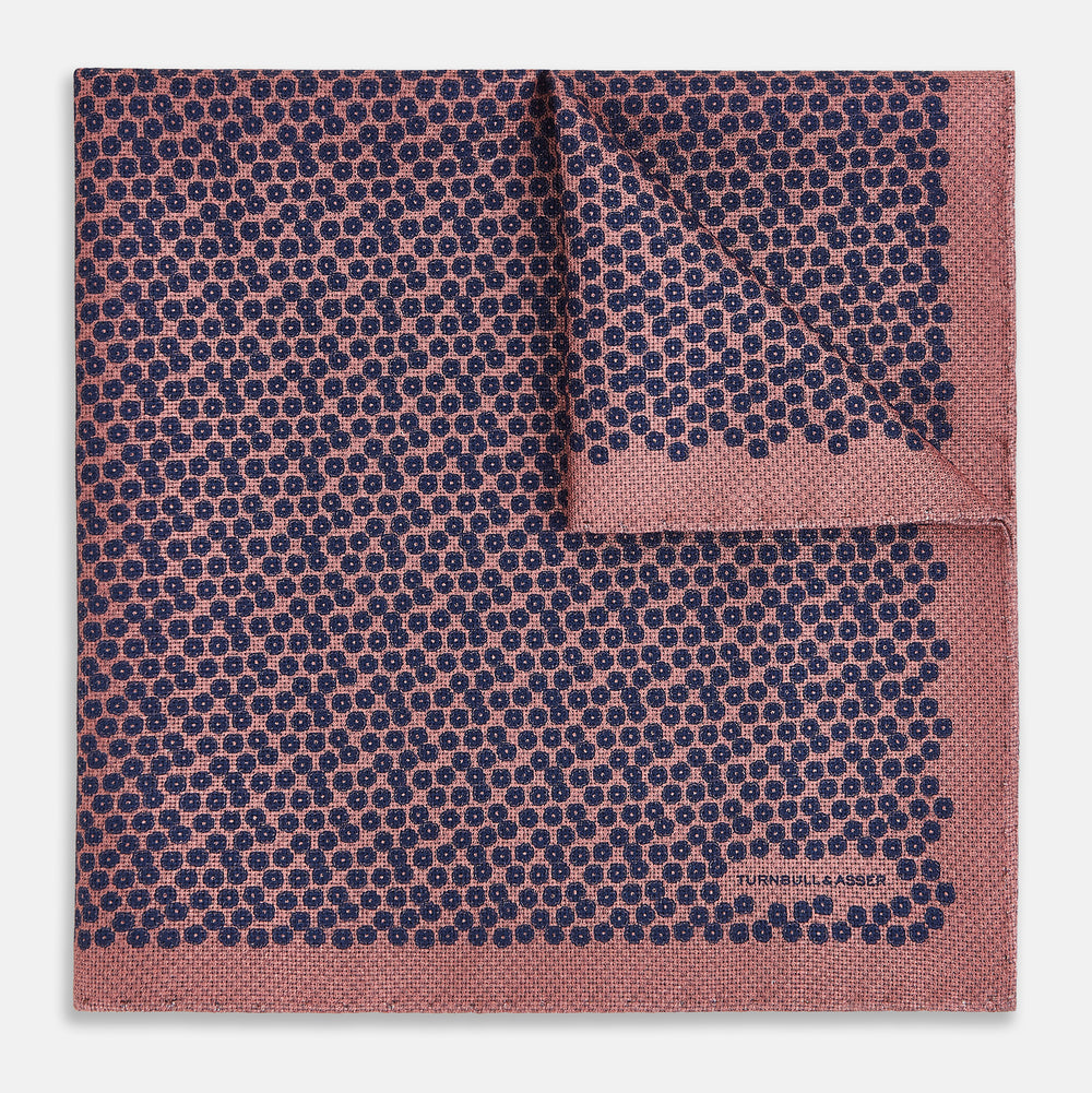 The Floral Pink Pocket Square in Silk by Turnbull & Asser UK showcases a refined pattern with a pink floral design and a contrasting border. The top right corner is elegantly folded to reveal the reverse side’s complementary blue and white dotted motif, emphasizing its double-sided versatility.