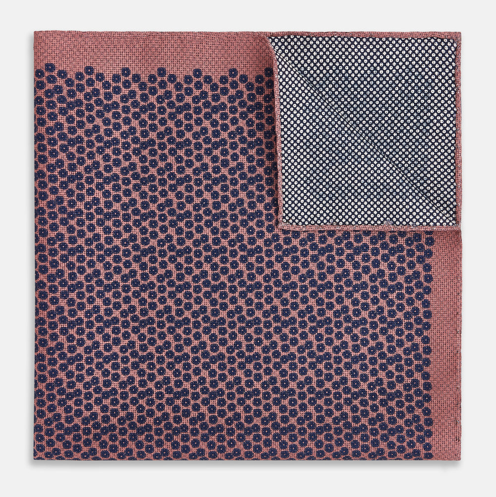 The Floral Pink Pocket Square in Silk by Turnbull & Asser UK showcases a refined pattern with a pink floral design and a contrasting border. The top right corner is elegantly folded to reveal the reverse side’s complementary blue and white dotted motif, emphasizing its double-sided versatility.