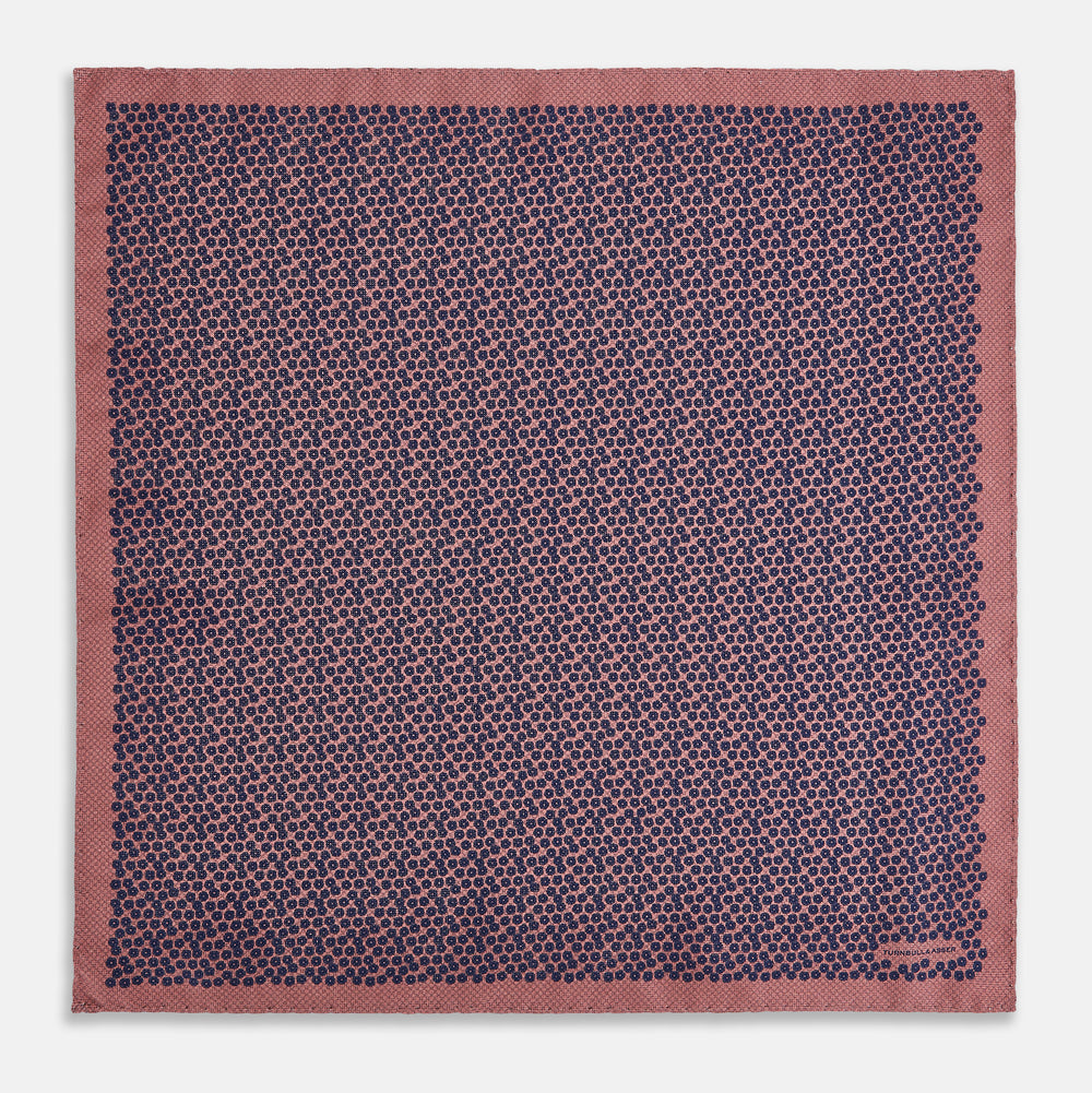 The Floral Pink Pocket Square in Silk by Turnbull & Asser UK showcases a refined pattern with a pink floral design and a contrasting border. The top right corner is elegantly folded to reveal the reverse side’s complementary blue and white dotted motif, emphasizing its double-sided versatility.