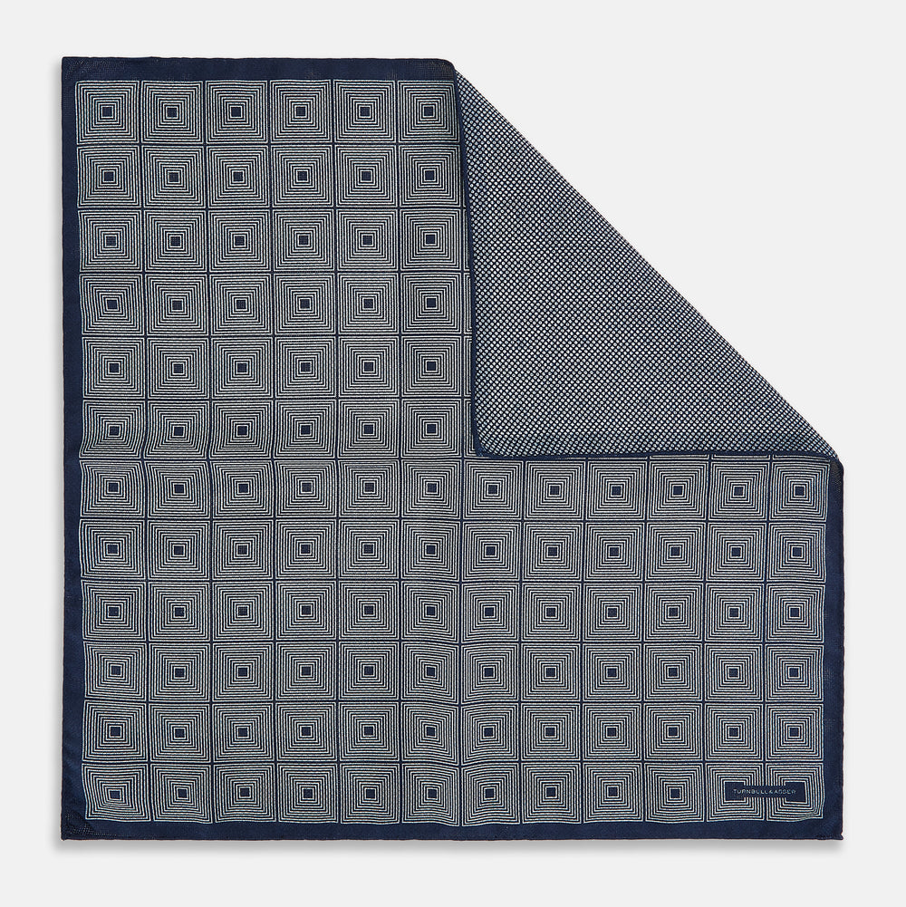 Introducing the Geometric Navy Tile Pocket Square in Silk by Turnbull & Asser UK—a folded navy and white accessory showcasing a double-sided design, with one side featuring concentric squares that form a striking geometric pattern and the other adorned with an elegant small dot motif.
