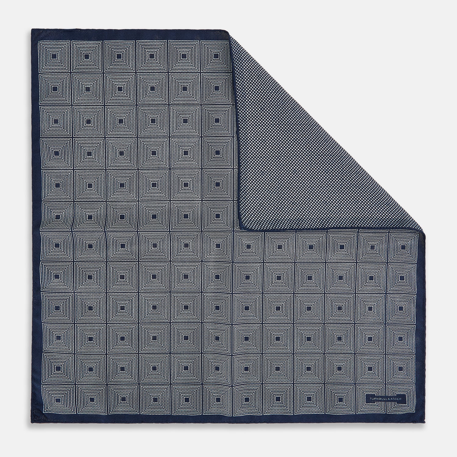 View 1 of product Introducing the Geometric Navy Tile Pocket Square in Silk by Turnbull & Asser UK—a folded navy and white accessory showcasing a double-sided design, with one side featuring concentric squares that form a striking geometric pattern and the other adorned with an elegant small dot motif.