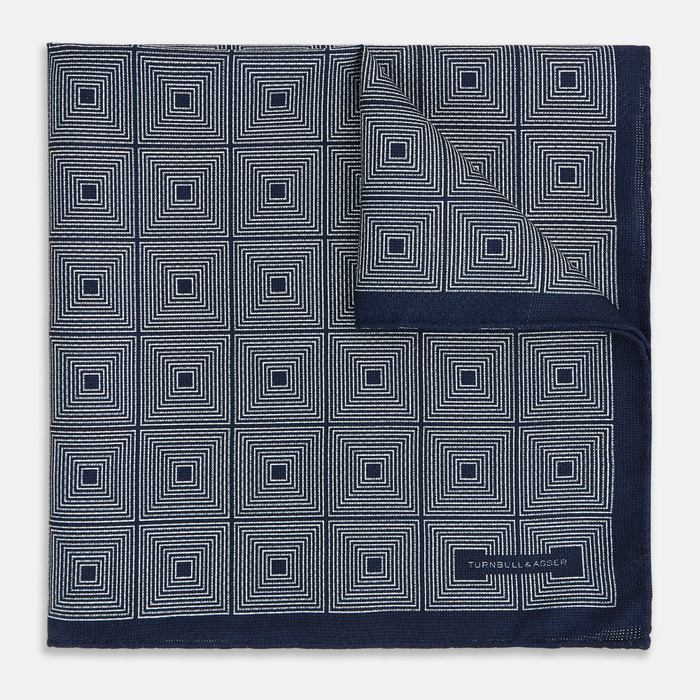 Introducing the Geometric Navy Tile Pocket Square in Silk by Turnbull & Asser UK—a folded navy and white accessory showcasing a double-sided design, with one side featuring concentric squares that form a striking geometric pattern and the other adorned with an elegant small dot motif.