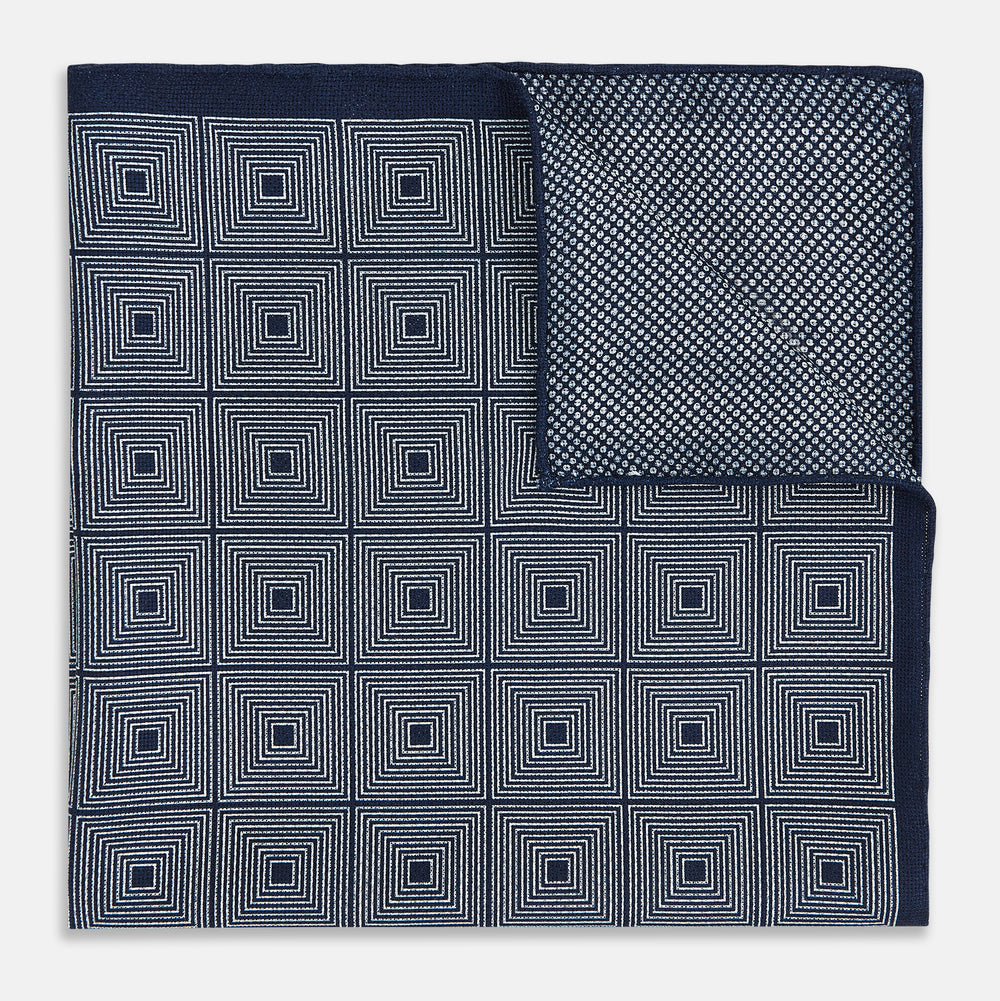 Introducing the Geometric Navy Tile Pocket Square in Silk by Turnbull & Asser UK—a folded navy and white accessory showcasing a double-sided design, with one side featuring concentric squares that form a striking geometric pattern and the other adorned with an elegant small dot motif.