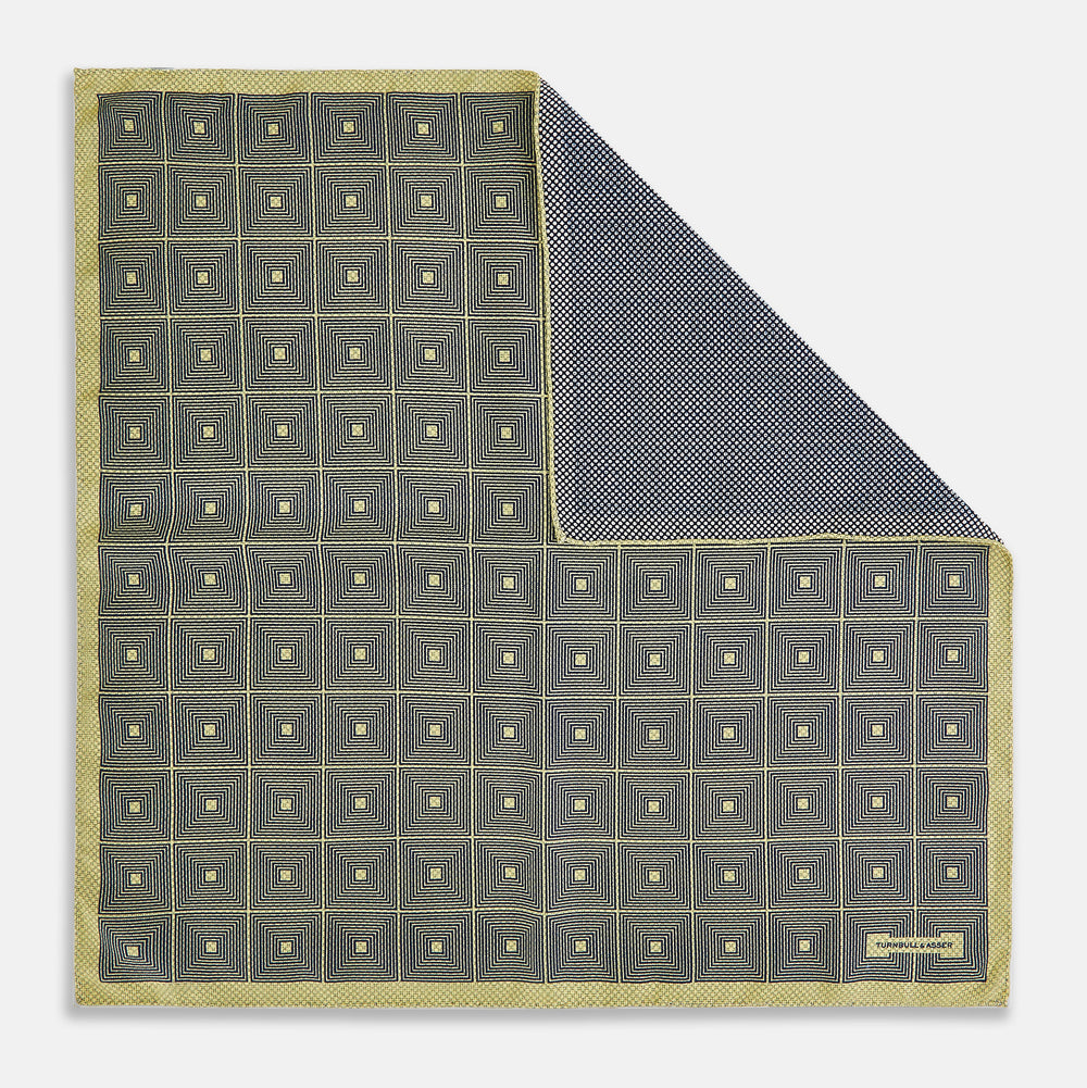 The Turnbull & Asser UK Geometric Yellow Tile Pocket Square in Silk features a sophisticated geometric square pattern in gold and navy, accented with subtle concentric squares and a discreet label stitched into the bottom right corner. Its double-sided design is highlighted by a folded corner that reveals a contrasting dotted navy and white underside.