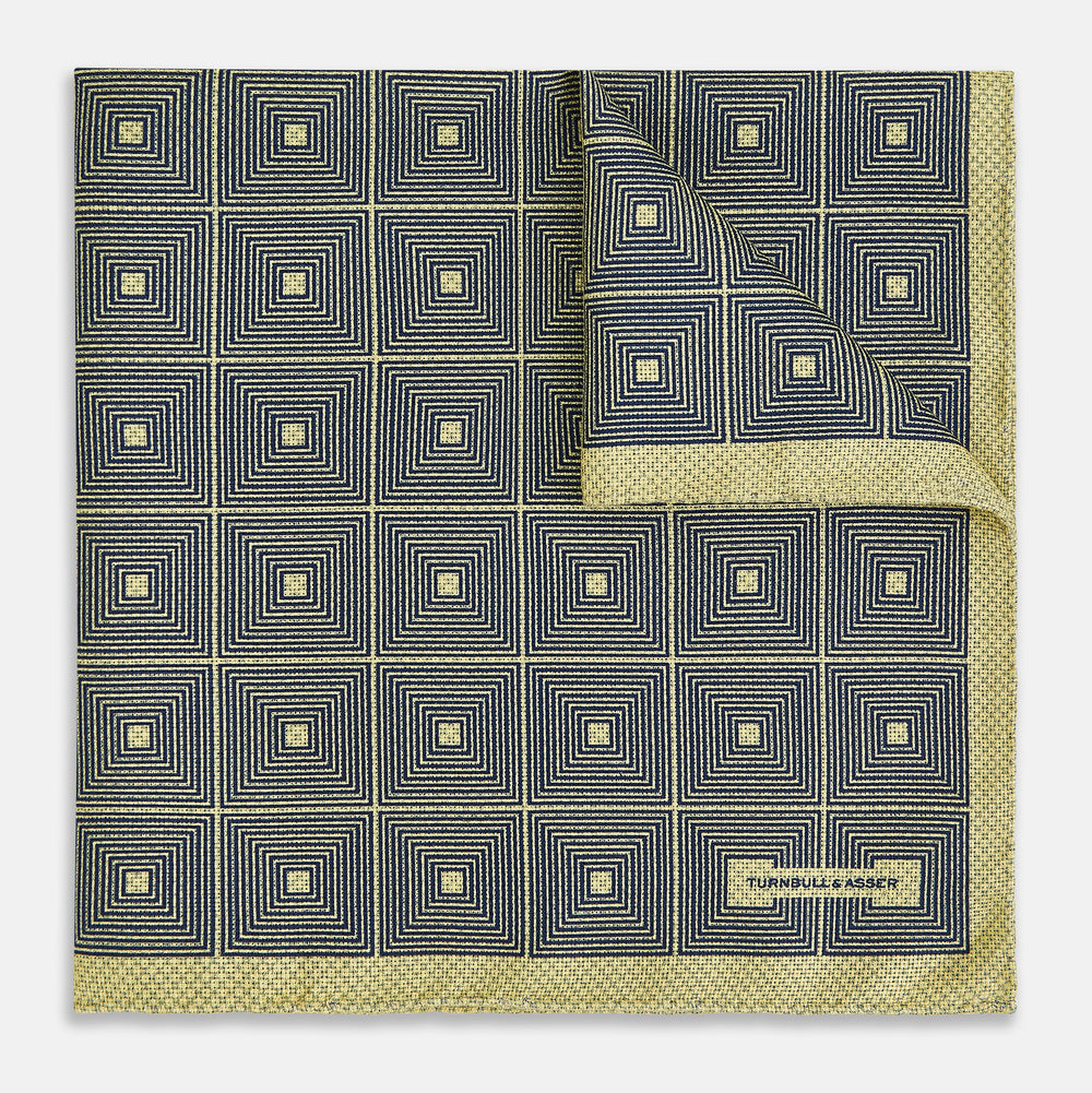 The Turnbull & Asser UK Geometric Yellow Tile Pocket Square in Silk features a sophisticated geometric square pattern in gold and navy, accented with subtle concentric squares and a discreet label stitched into the bottom right corner. Its double-sided design is highlighted by a folded corner that reveals a contrasting dotted navy and white underside.