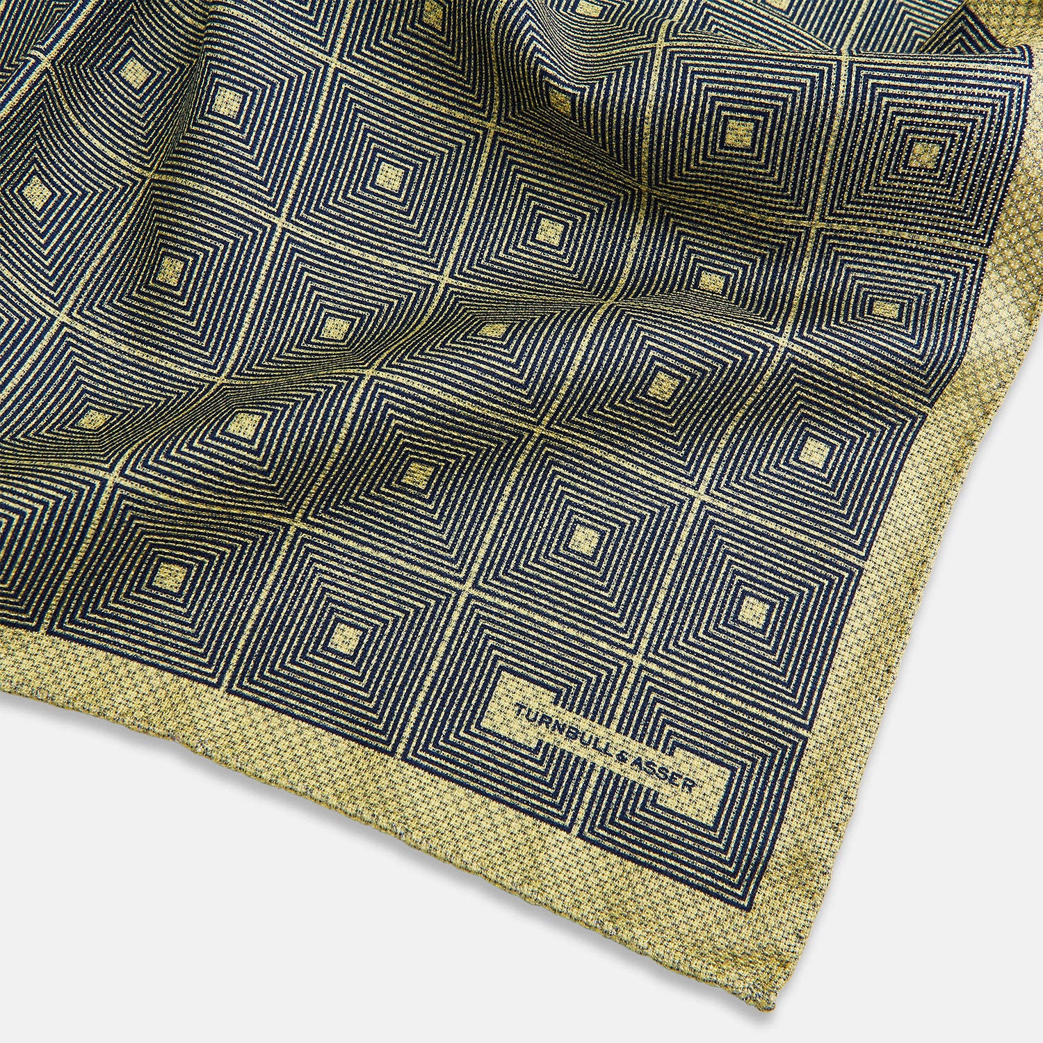 View 3 of product The Turnbull & Asser UK Geometric Yellow Tile Pocket Square in Silk features a sophisticated geometric square pattern in gold and navy, accented with subtle concentric squares and a discreet label stitched into the bottom right corner. Its double-sided design is highlighted by a folded corner that reveals a contrasting dotted navy and white underside.