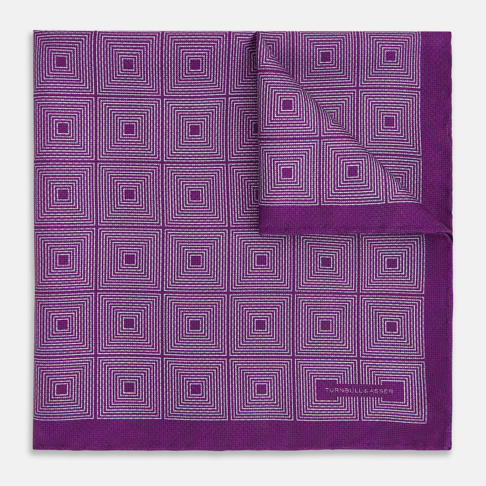 The Turnbull & Asser UK Geometric Purple Tile Pocket Square in Silk features a vibrant purple hue adorned with concentric square motifs. One corner is folded to reveal its double-sided design, showcasing a contrasting black-and-white dotted underside.