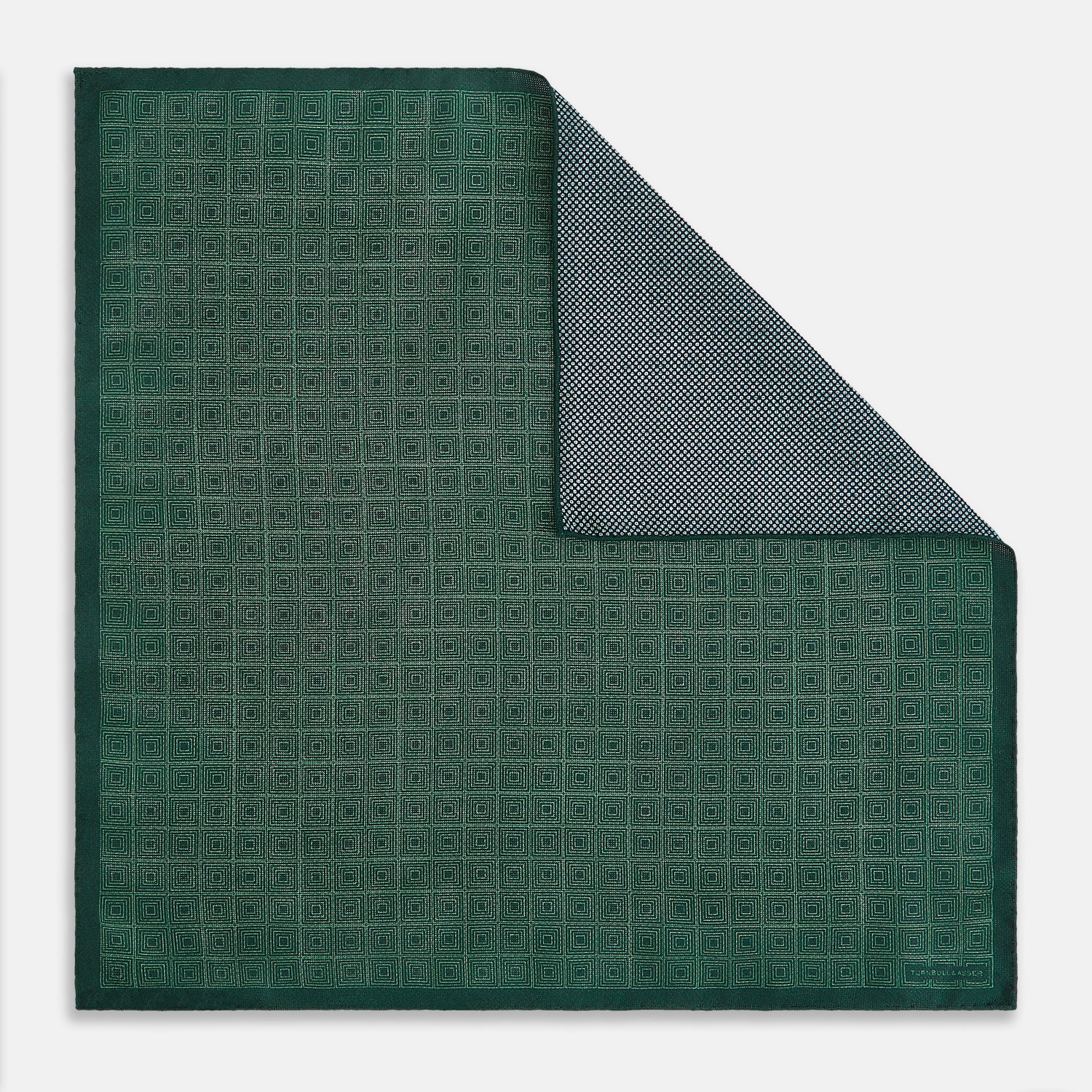 View 1 of product The Geometric Green Tile Pocket Square in Silk by Turnbull & Asser UK features a sophisticated tonal green design with a subtle pattern of concentric squares. One corner is folded to highlight the elegant double-sided construction, revealing a contrasting black and white small dot pattern on the reverse.