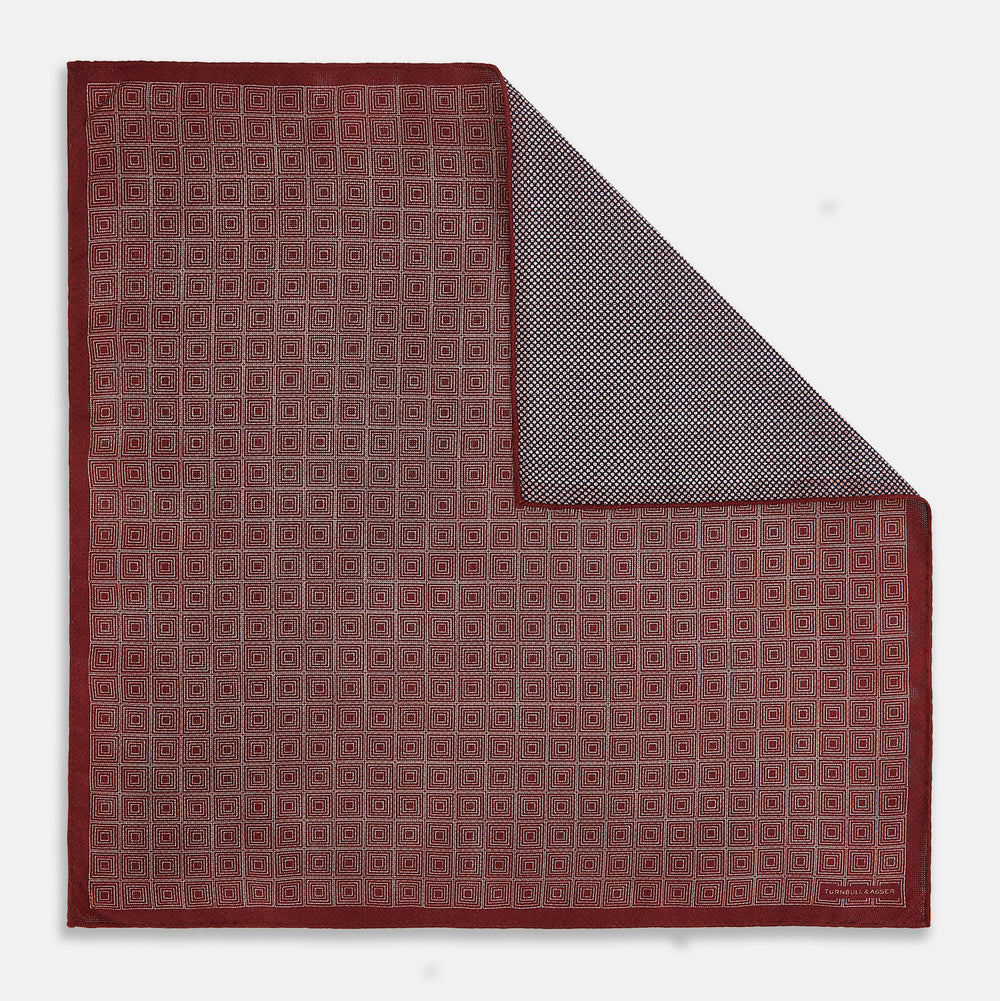 The Turnbull & Asser UK Geometric Burgundy Tile Pocket Square in Silk is displayed partly folded, showcasing its bold geometric grid pattern with concentric burgundy squares on the front and a contrasting black-and-white dotted design on the reverse.