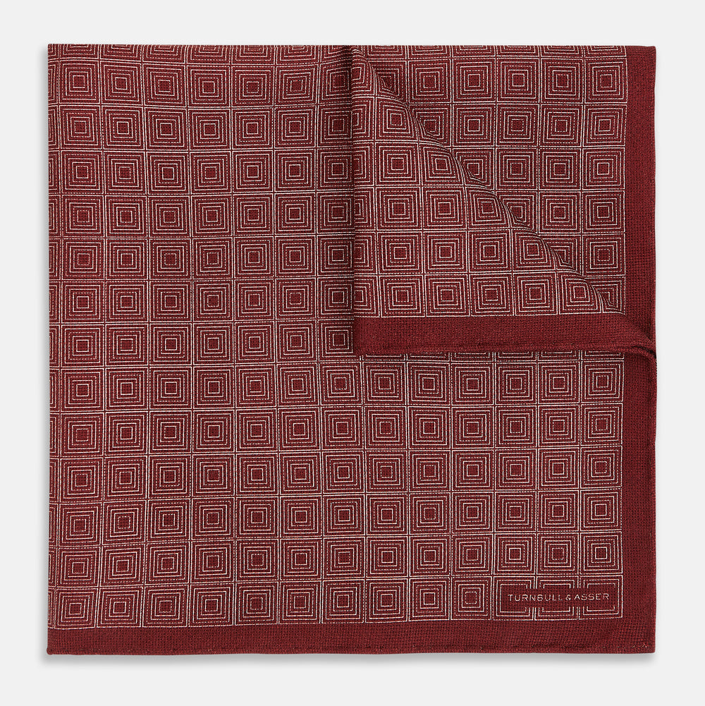 The Turnbull & Asser UK Geometric Burgundy Tile Pocket Square in Silk is displayed partly folded, showcasing its bold geometric grid pattern with concentric burgundy squares on the front and a contrasting black-and-white dotted design on the reverse.
