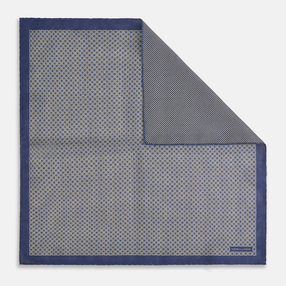 The Geometric Blue Pocket Square in Silk from Turnbull & Asser UK showcases a blue border and double-sided design, with a geometric pattern at the center. A folded corner reveals a refined, tighter dot motif on the reverse side, emphasizing the intricate craftsmanship of this finely woven accessory.