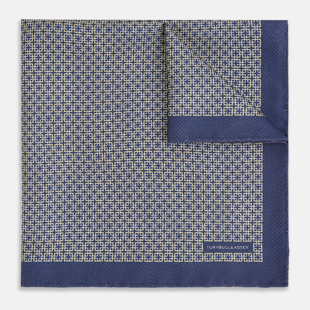 The Geometric Blue Pocket Square in Silk from Turnbull & Asser UK showcases a blue border and double-sided design, with a geometric pattern at the center. A folded corner reveals a refined, tighter dot motif on the reverse side, emphasizing the intricate craftsmanship of this finely woven accessory.