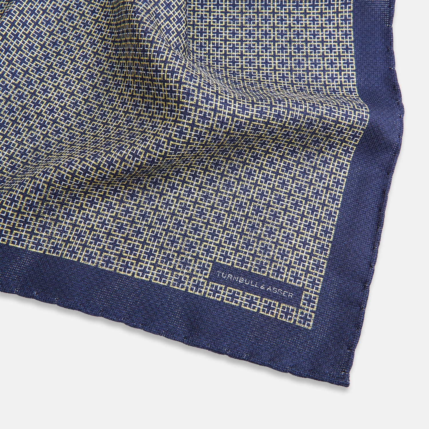 View 3 of product The Geometric Blue Pocket Square in Silk from Turnbull & Asser UK showcases a blue border and double-sided design, with a geometric pattern at the center. A folded corner reveals a refined, tighter dot motif on the reverse side, emphasizing the intricate craftsmanship of this finely woven accessory.