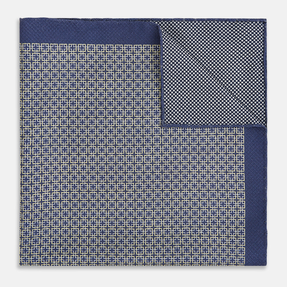 The Geometric Blue Pocket Square in Silk from Turnbull & Asser UK showcases a blue border and double-sided design, with a geometric pattern at the center. A folded corner reveals a refined, tighter dot motif on the reverse side, emphasizing the intricate craftsmanship of this finely woven accessory.