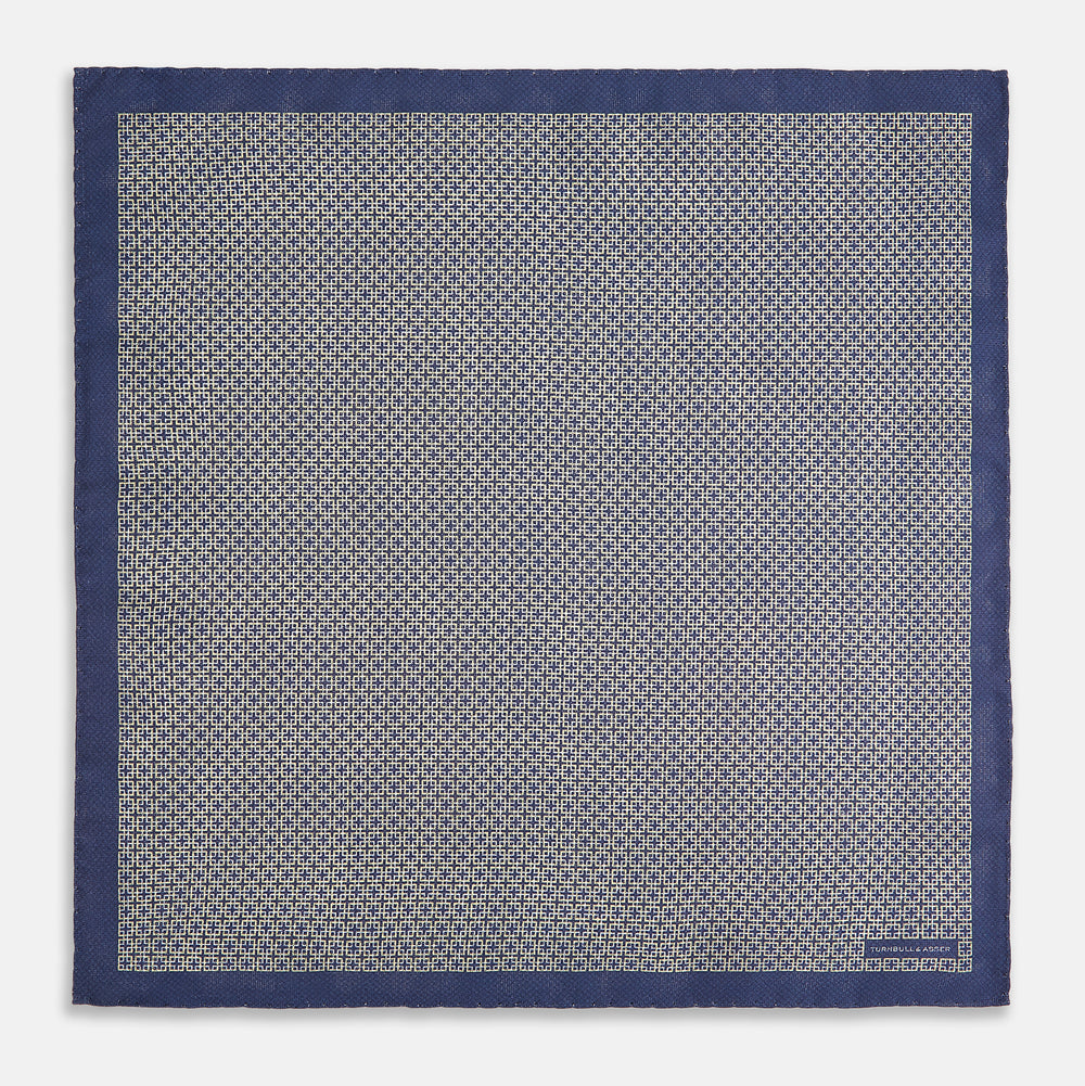 The Geometric Blue Pocket Square in Silk from Turnbull & Asser UK showcases a blue border and double-sided design, with a geometric pattern at the center. A folded corner reveals a refined, tighter dot motif on the reverse side, emphasizing the intricate craftsmanship of this finely woven accessory.