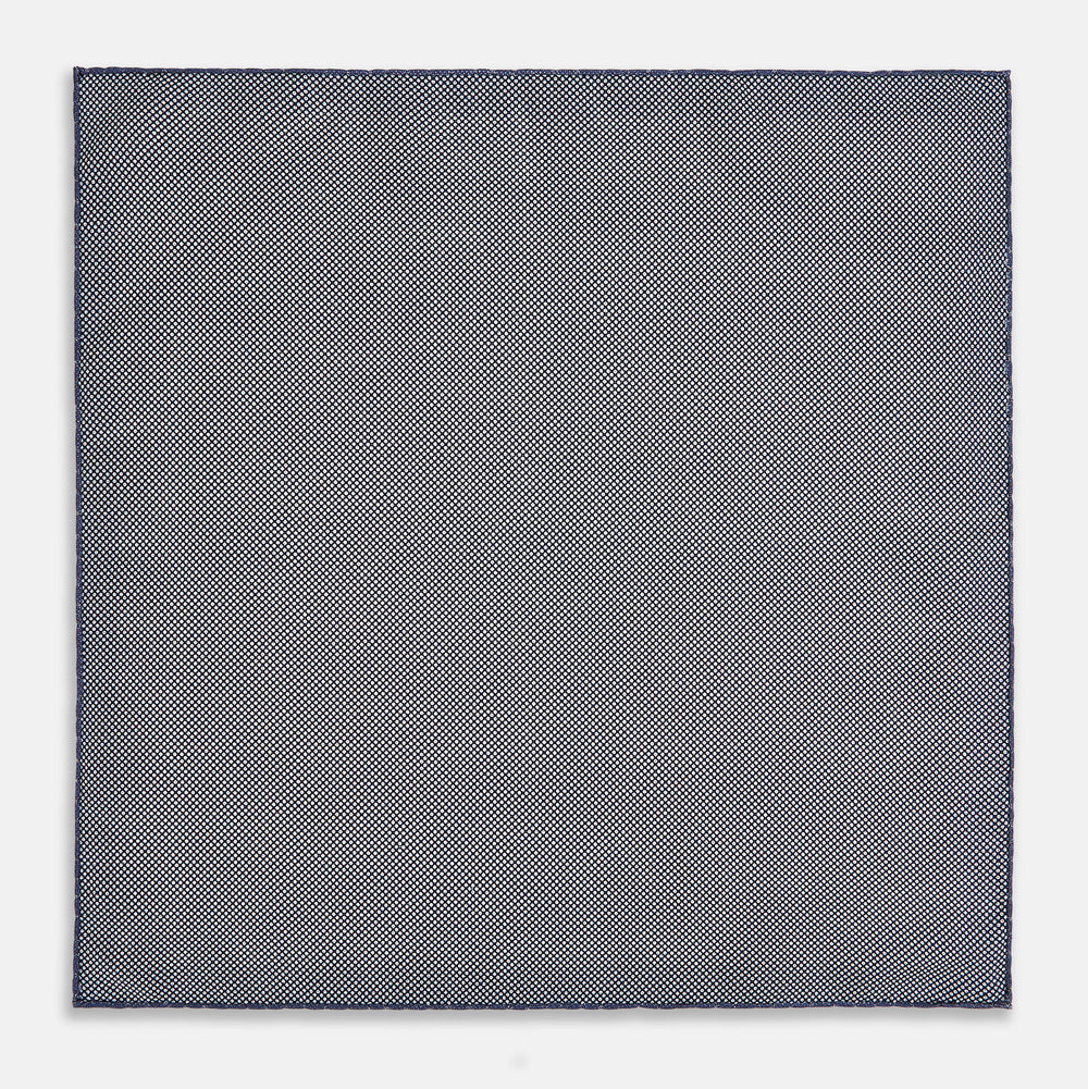 The Geometric Blue Pocket Square in Silk from Turnbull & Asser UK showcases a blue border and double-sided design, with a geometric pattern at the center. A folded corner reveals a refined, tighter dot motif on the reverse side, emphasizing the intricate craftsmanship of this finely woven accessory.