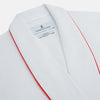 View 2 of product The Turnbull & Asser White Piped Herringbone Cotton Gown features long sleeves, a waist tie, chest pocket, and striking red piping on the collar, cuffs, pocket, and edges—an elegant piece showcased against a white background.