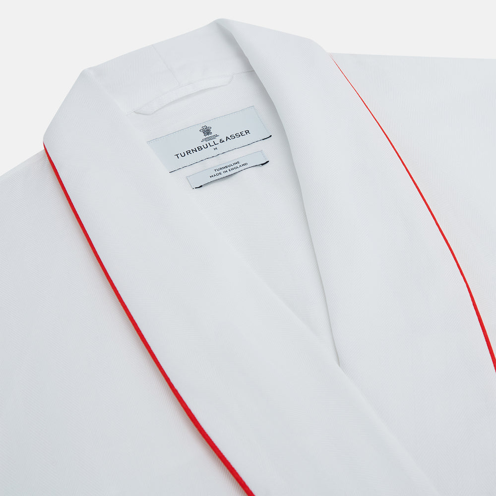 The Turnbull & Asser White Piped Herringbone Cotton Gown features long sleeves, a waist tie, chest pocket, and striking red piping on the collar, cuffs, pocket, and edges—an elegant piece showcased against a white background.