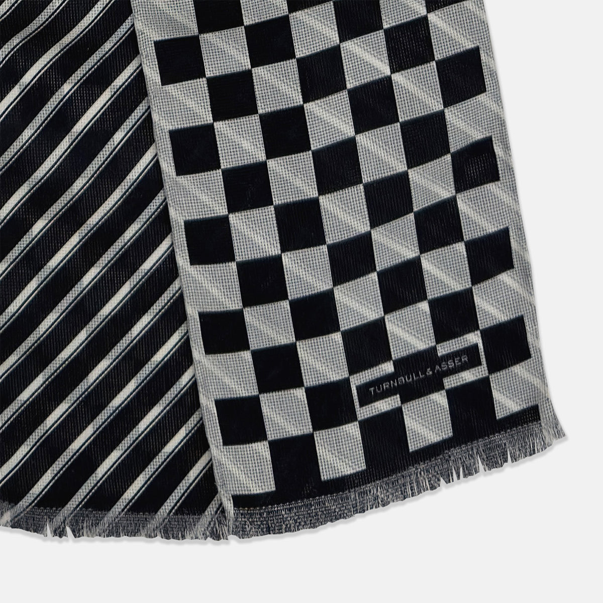 View 3 of product The Chequered Navy on White Frame Small Scarf in Silk by Turnbull & Asser UK is a long silk accessory featuring a classic black and white checkerboard pattern laid flat against a white background. Its ends are finished with a thin, fringed border, while the top edge is accented by diagonal stripes. The double-sided chequered design adds a stylish twist, with touches of navy and subtle hints of white bringing sophisticated detail to this timeless look.