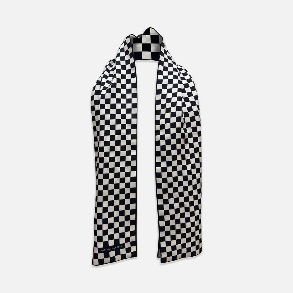 A Chequered Navy & White Scarf in Silk by Turnbull & Asser UK is displayed against a plain white background, showcasing its long, luxurious silk fabric with a striking navy and white checkered pattern.