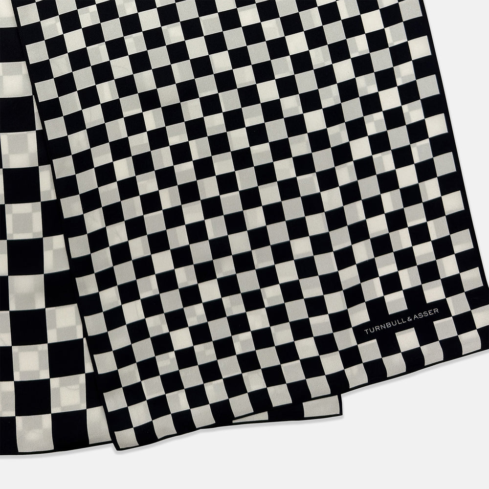 A Chequered Navy & White Scarf in Silk by Turnbull & Asser UK is displayed against a plain white background, showcasing its long, luxurious silk fabric with a striking navy and white checkered pattern.