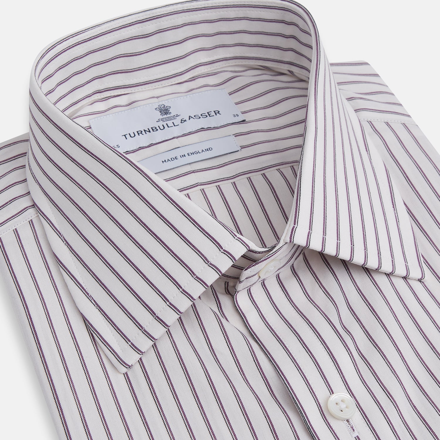 View 2 of product The Turnbull & Asser Purple Track Stripe Mayfair Shirt is a long-sleeve, button-up organic cotton shirt with a T&A collar, showcasing thin vertical black stripes on white and laid flat on a white surface.