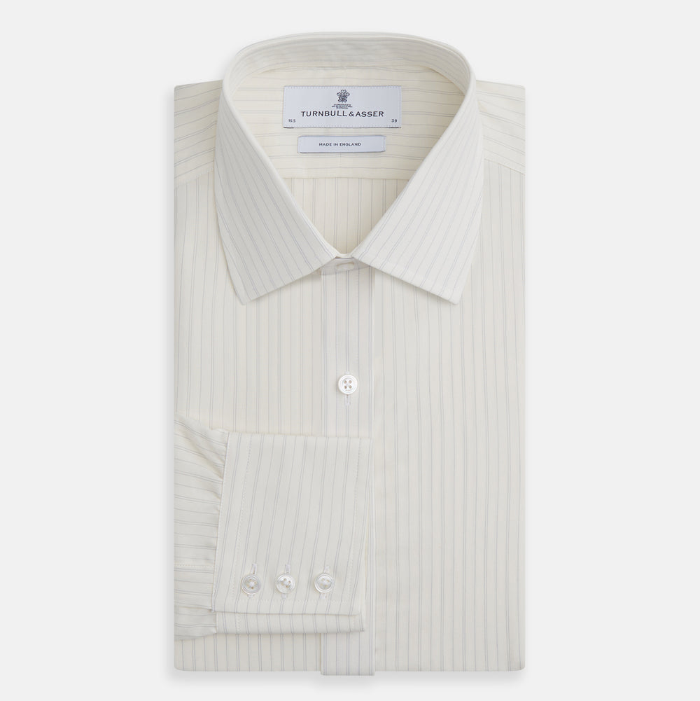 The Turnbull & Asser Pale Blue Fine Track Stripe Mayfair Shirt is a long-sleeve, cream-colored organic cotton shirt with subtle pale blue stripes, a T&A collar, and buttoned cuffs, shown on a plain white background.