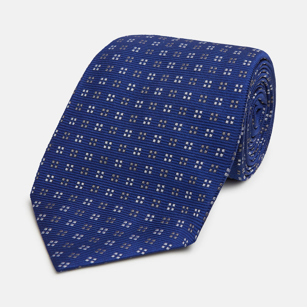 The Turnbull & Asser Blue and Brown Multi Dot Silk Tie, shown rolled up on a plain white background, features an elegant design with small white and gray dots arranged in squares.