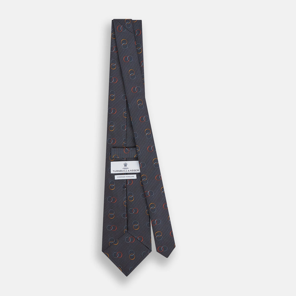The Navy Links Silk Tie by Turnbull & Asser, featuring a pattern of overlapping red, yellow, and blue rings, is rolled up and displayed against a plain white background.