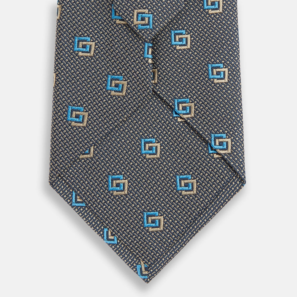 The Turnbull & Asser Blue and Gold Silk Tie, featuring a refined repeating geometric pattern, exudes British elegance and is displayed rolled up on a plain white background.