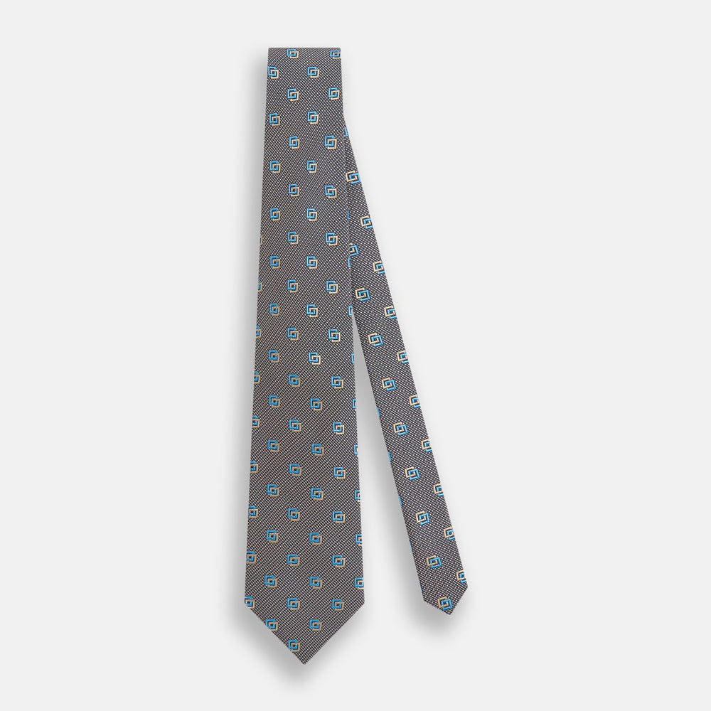 The Turnbull & Asser Blue and Gold Silk Tie, featuring a refined repeating geometric pattern, exudes British elegance and is displayed rolled up on a plain white background.