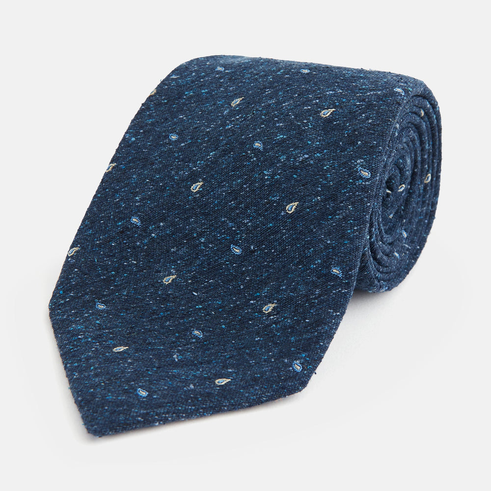The Turnbull & Asser Navy Micro Paisley Silk Tie features a rolled-up navy blue silk with a subtle speckled texture and small light blue paisley patterns, perfect for enhancing any formal collection, shown on a white background.