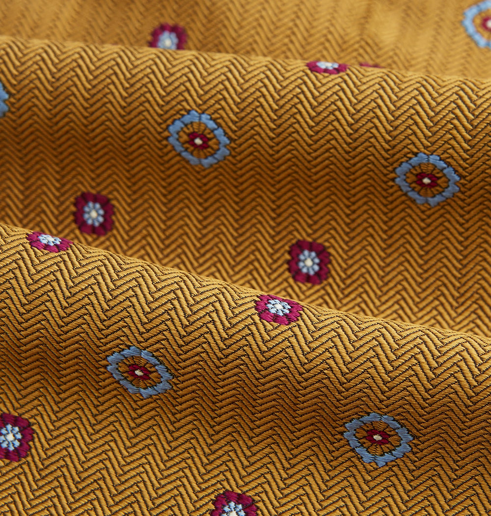 The Turnbull & Asser Gold Motif Silk Cravat features a herringbone pattern with blue and red floral accents—stylishly folded with a pointed tip, it's ideal for elevating your look at formal events.