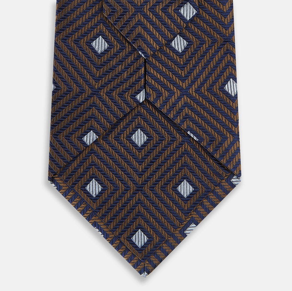 The Tomorrow Never Dies Seven-Fold Silk Tie by Turnbull & Asser UK features a geometric pattern of navy and brown squares with light blue diamond accents, rolled up and displayed on a plain white background.