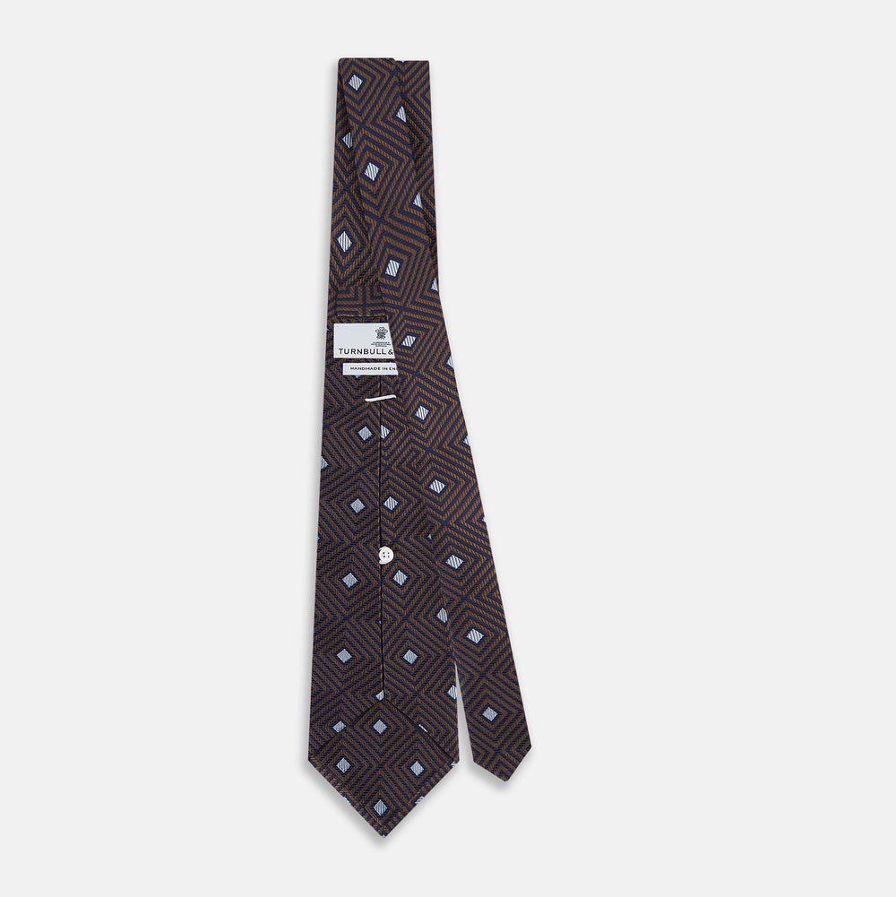 The Tomorrow Never Dies Seven-Fold Silk Tie by Turnbull & Asser UK features a geometric pattern of navy and brown squares with light blue diamond accents, rolled up and displayed on a plain white background.
